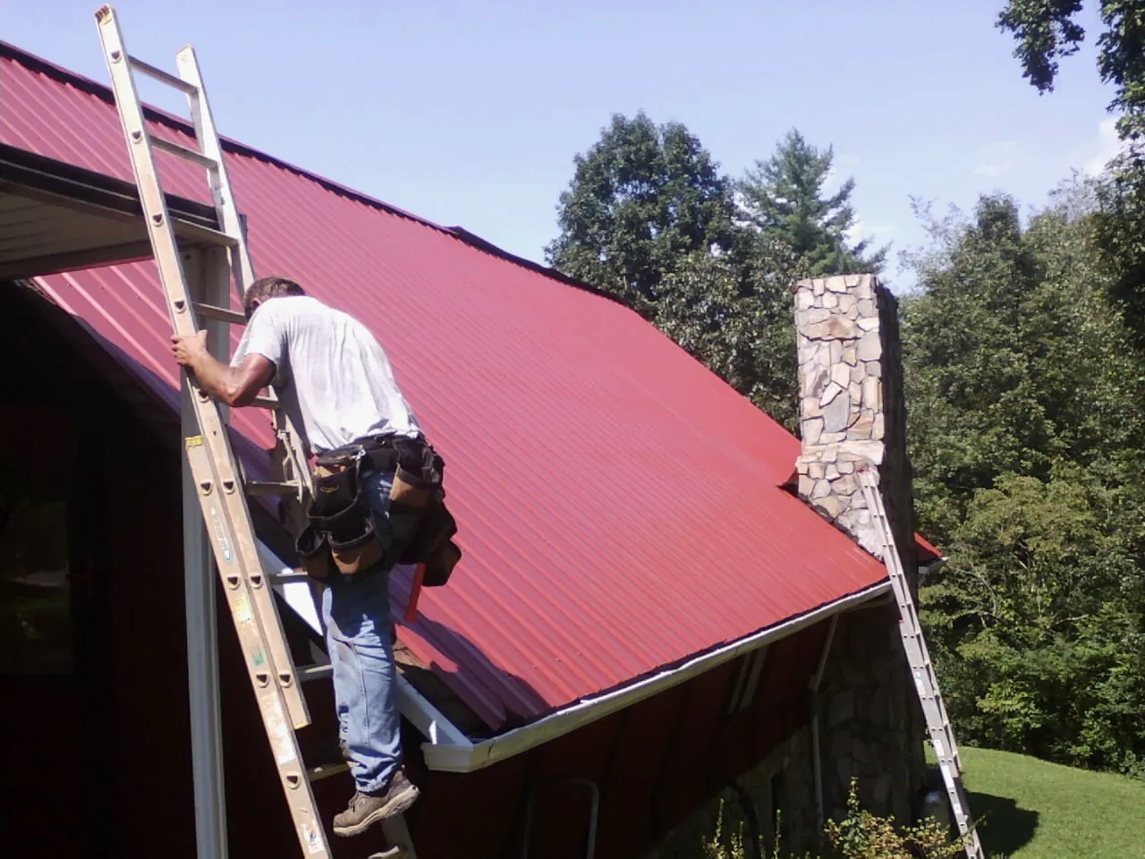 Roofer on a ladder beside a red metal roof and stone chimney — Soffit & Fascia Repair by Roofing Brothers in Jordan